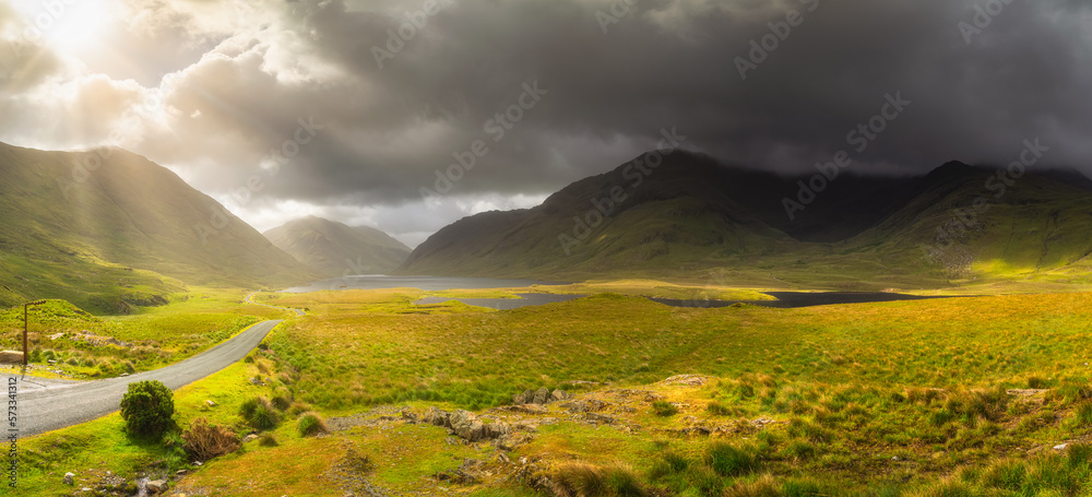 Panorama of Doolough Valley with lakes, Glenummera and Glencullin mountain ranges illuminated by sunlight, dark dramatic sky, County Mayo, Ireland