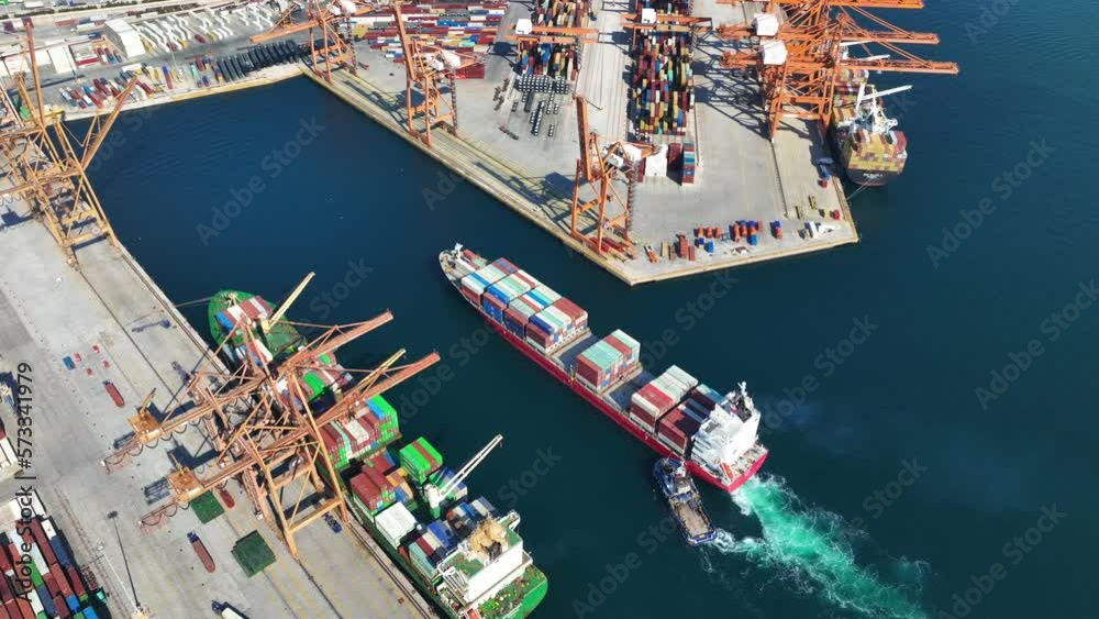 Aerial top down video of industrial cargo container ship being towed to logistics container terminal port to dock