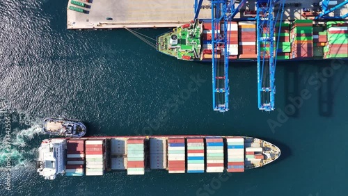 Aerial top down video of industrial cargo container ship being towed to logistics container terminal port to dock