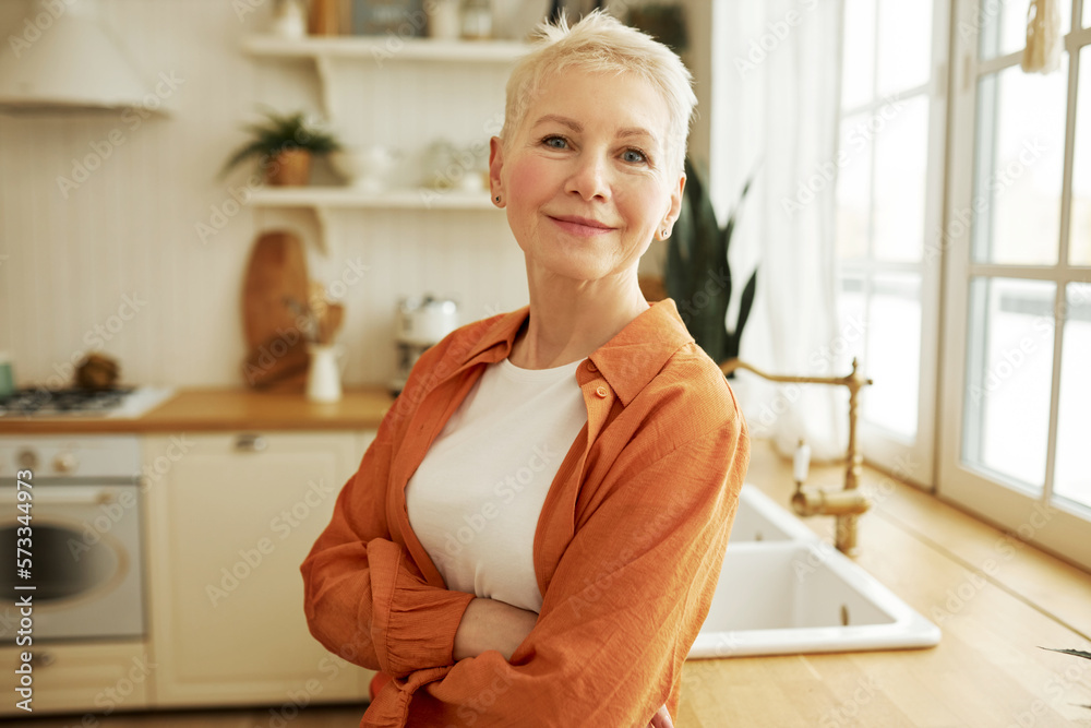Indoor image of attractive happy confident mature female house owner standing in kitchen leaning against counter with big window, looking at camera with crossed hands and positive facial expression Indoor image of attractive happy confident mature female house owner standing in kitchen leaning against counter with big window, looking at camera with crossed hands and positive facial expression
