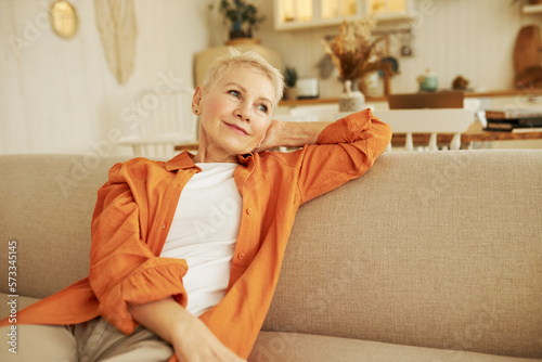 Happy mature elderly caucasian woman relaxing on couch, enjoying weekend or leisure time, having rest after homework on sofa, looking aside with positive facial expression and smile