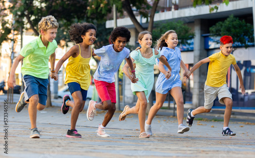 Fototapeta Naklejka Na Ścianę i Meble -  Group of cheerful tweenagers of different nationalities running together along city street on summer day. Happy healthy kids concept..