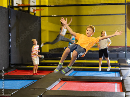 Full length of excited casual schoolchild boy having great time while jumping high on colorful trampoline at game club