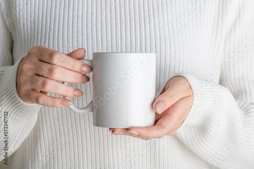 Female hands holding white mug mockup with blank copy space for your advertising text message or promotional content. Girl in white sweater holding white porcelain coffee mug mock up, close up