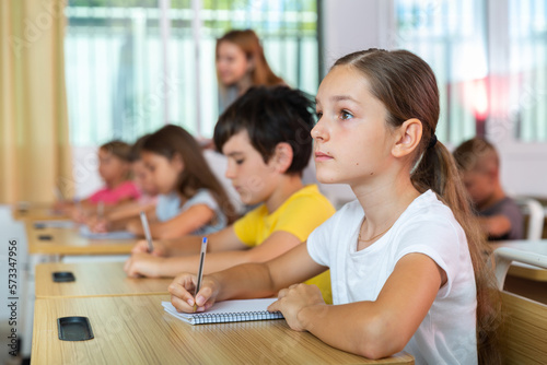 Portrait of focused preteen schoolgirl writing exercises in workbook during lesson in classroom ..