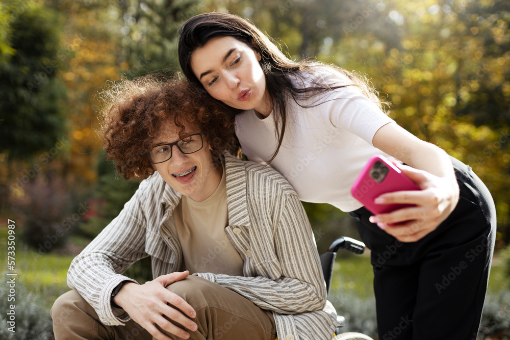 Fototapeta premium Positive, young man in a wheelchair and his girlfriend having fun outdoors while taking a selfie. Young people make funny spades.
