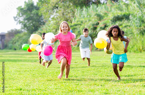 Happy tween friends of different nationalities having fun together in summer city park, chasing each other on green meadow, holding colorful balloons in hands..