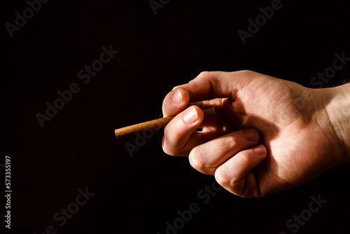 A man's hand holds a lit cigarette on a dark background. Hard light. Smoking. Bad habits of people. Lungs' cancer. Fight against smoking.