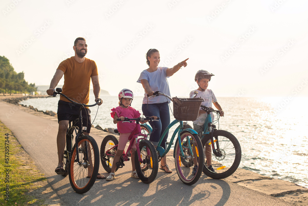 Fototapeta premium Young family, parents with two children, on bikes standing near the sea, looking at view in the direction that the mother pointing