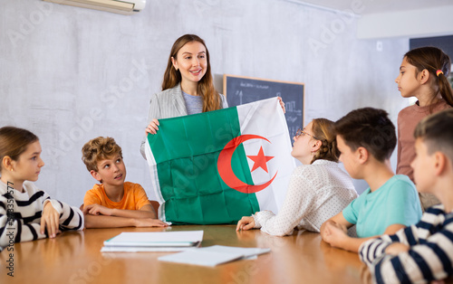 Decent pedagogue showing Algeria flag to group of preteen schoolchildren in classroom during lesson
