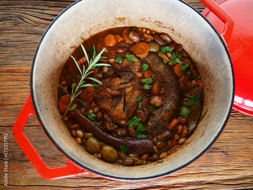French Cassoulet, a meat and beans stew, in a red cast iron cooking pot, on a rustic wooden table