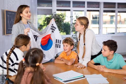 Happy young woman teacher showing national flag of South Korea and telling preteens schoolchildren history of country during lesson in class