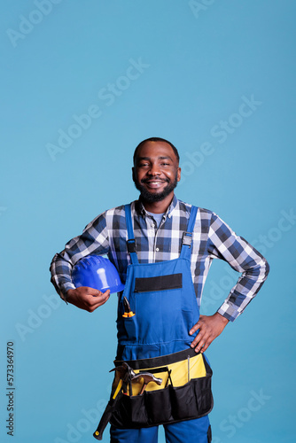 African american construction worker looking proud at work place holding hard hat. Smiling handyman wearing coveralls and tool belt isolated on blue background, studio shot. © DC Studio