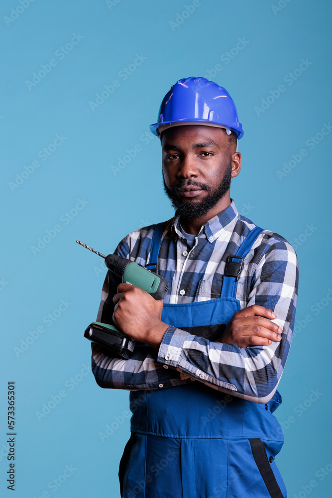 African american builder holds electric cordless drill while looking seriously into camera. Contractor holding work tool with confidence, expert in construction and renovations.