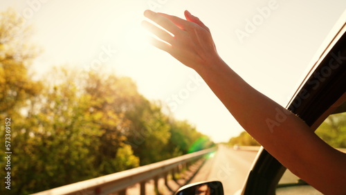 Free young girl waves her hand from car window, travels, catches sun glare with her fingers. Auto travel on road on vacation. Young woman driver plays catches fresh wind with hand from car window.