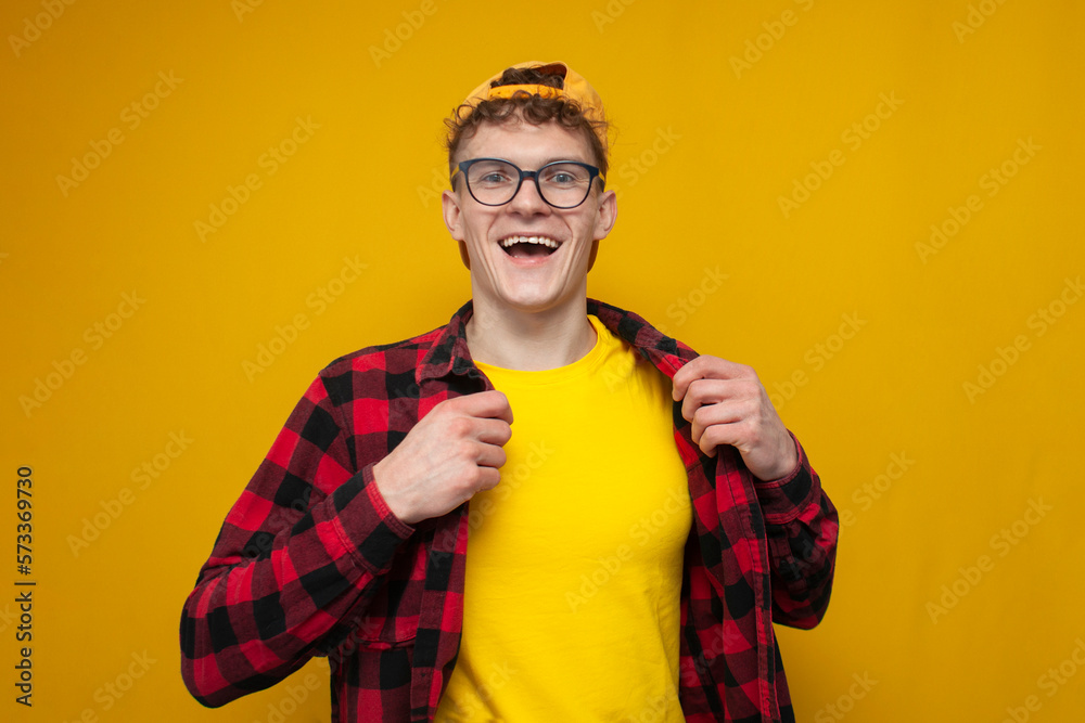 confident guy in glasses with curly hair puts on a shirt on a yellow background