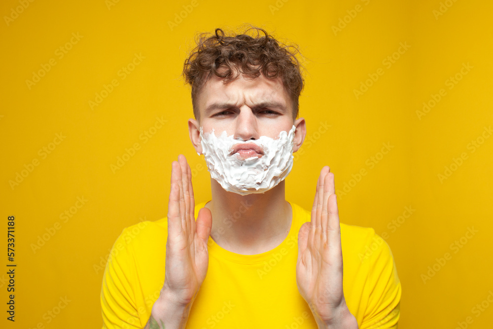 curly guy with shaving foam on his face is surprised and looks away on a yellow background, student shaves his beard