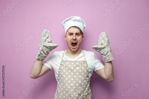 shocked baker in uniform with gloves raises his hands up and shouts, chef in apron in stress photo