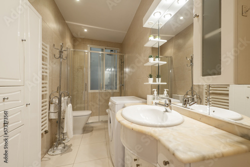 Bathroom with cream marble sink with wooden cabinet with integrated mirror with lights and shelves and washing machine in one corner