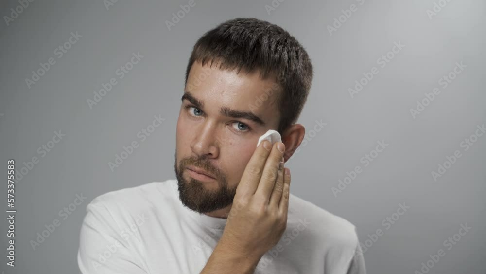 Handsome man is cleaning his face with a cotton pad on gray background Handsome man is cleaning his face with a cotton pad on gray background
