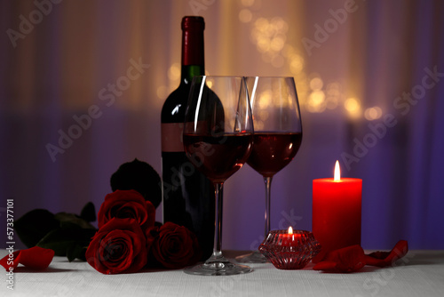Glasses of red wine, rose flowers and burning candles on grey table against blurred lights. Romantic atmosphere