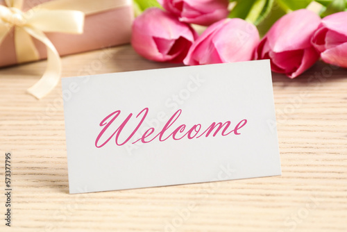 Welcome card, gift box and beautiful pink tulips on wooden table, closeup
