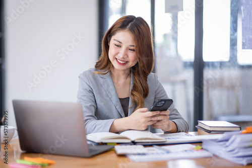 Smiling young Asian business woman executive looking at smartphone using cellphone mobile cell tech  happy ethnic professional female worker working in office typing on cellphone sitting at desk.