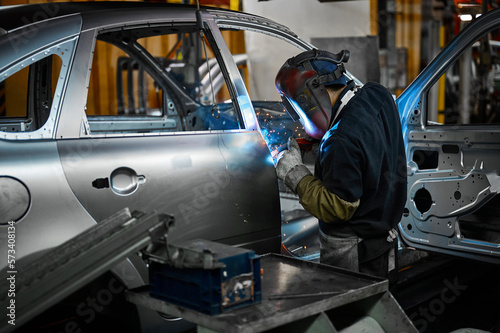 Fotografie Worker in mask welds metal parts of auto carcass in shop