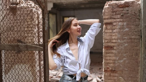 Attractive brown-haired young woman in blue men shirt, tied in knot on tummy, slightly revealing breasts, posing near old rusty wire fencing in dilapidated abandoned house