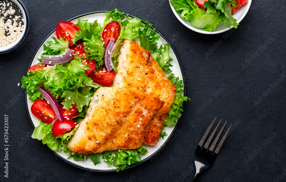 Fried cod fish with salad garnish from lettuce, cherry tomatoes and red onion with sesame seeds, black table background, top view Fried cod fish with salad garnish from lettuce, cherry tomatoes and red onion with sesame seeds, black table background, top view