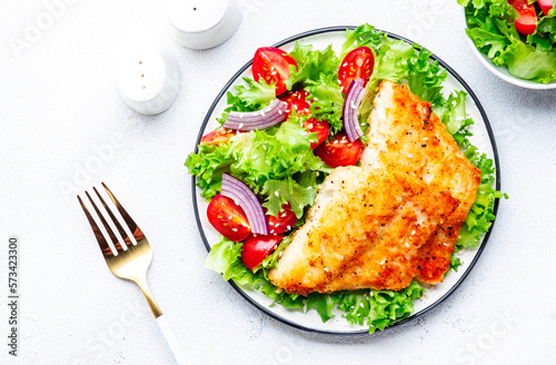Fried cod fish with salad garnish from lettuce, cherry tomatoes and red onion with sesame seeds, white table background, top view