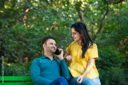 Young indian man sitting with young woman at park and talking on smartphone.