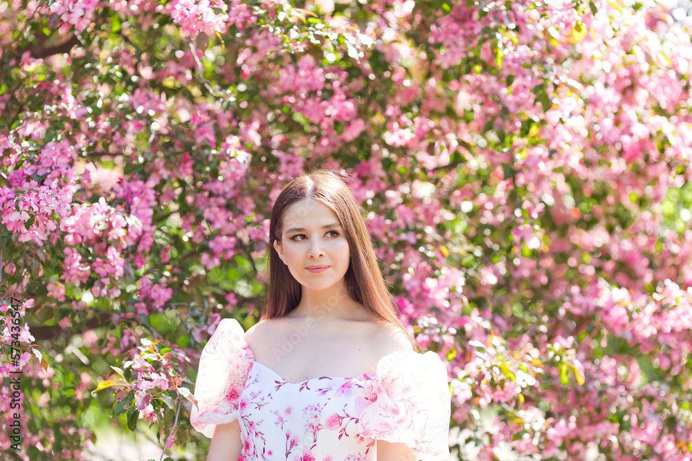 Portrait of brunette girl in pink dress, is standing under a pink blooming apple trees