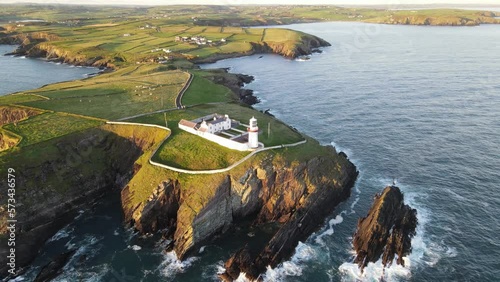 Stunning aerial footage captures Irish Lighthouse Galley Head On The Coast Of Co. Cork surrounded by cliffs, sun kissed rolling waves and rocky cliffs all at golden hour. photo