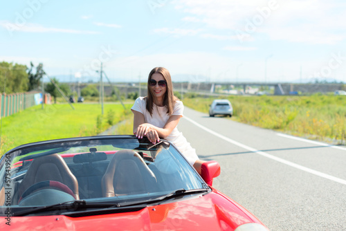 Lifestyle portrait of a carefree beautiful woman in white dress and sunglasses sitting on red cabriolet car and smiling. Road trip enjoying freedom concept  © Rina Mskaya