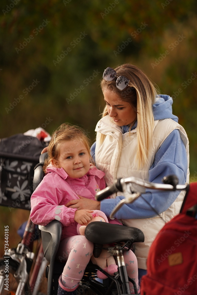 Portrait of little toddler girl sitting in bike seat and her mother with bicycle. Safe and child protection concept. Family and weekend activity trip