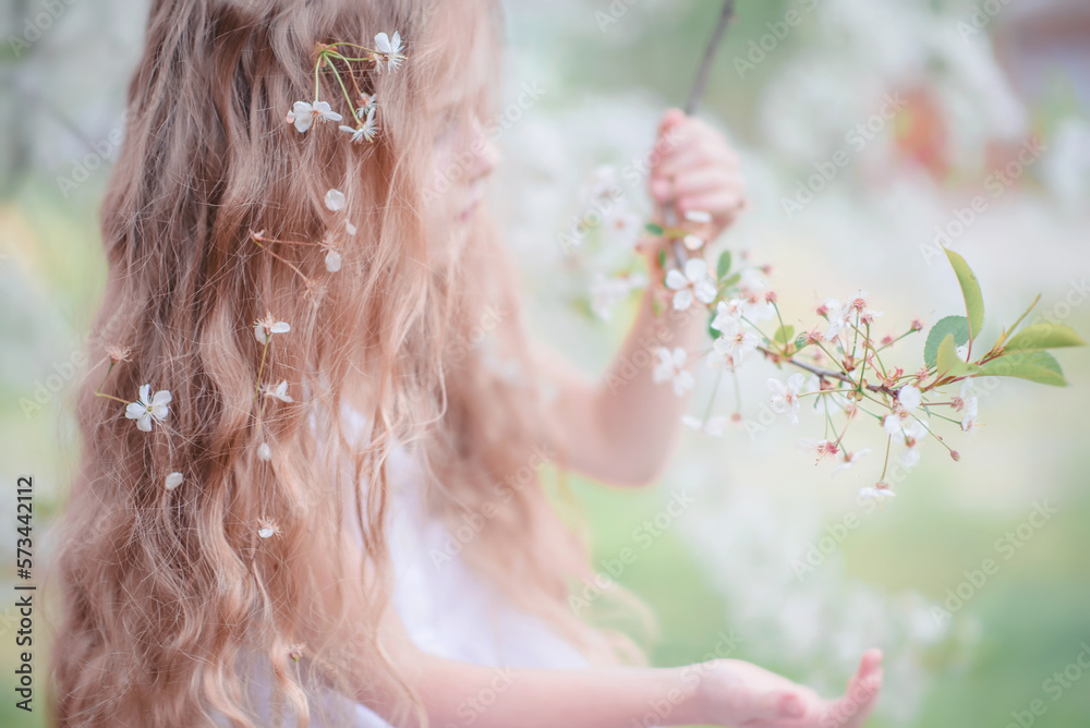 Naklejka premium Spring. A beautiful little girl with long hair and flowers in her hair near a flowering tree. Apple tree in bloom. The Cherry Orchard. Apple orchard. The baby is holding a twig. Portrait. tender baby