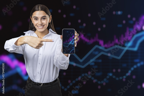 Fototapeta Naklejka Na Ścianę i Meble -  Attractive happy young european woman pointing at cellphone with forex chart on dark background. Worker, success, market, trade and invest concept.