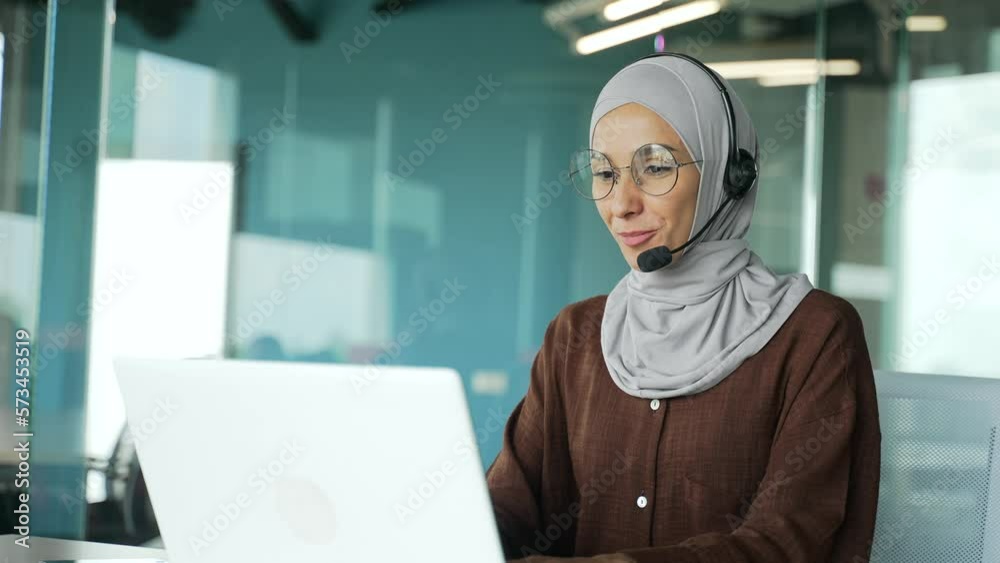 Positive young muslim businesswoman support manager in hijab with wireless headset looking at laptop screen camera and talking by online meeting conference video call at workplace in modern office Positive young muslim businesswoman support manager in hijab with wireless headset looking at laptop screen camera and talking by online meeting conference video call at workplace in modern office
