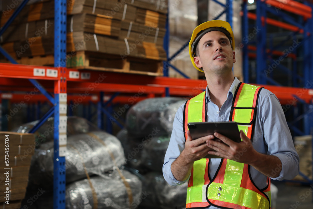 Warehouse workers man with hardhats and reflective jackets using tablet, walkie talkie radio and cardboard while controlling stock and inventory in retail warehouse logistics, distribution center Warehouse workers man with hardhats and reflective jackets using tablet, walkie talkie radio and cardboard while controlling stock and inventory in retail warehouse logistics, distribution center