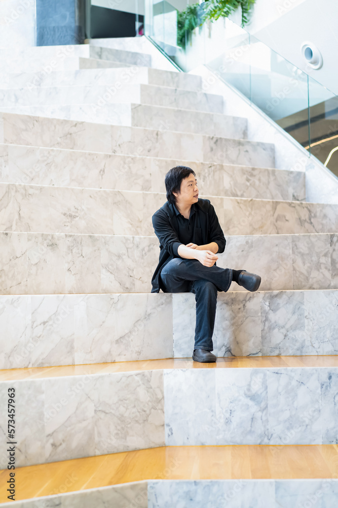 Asian Man in Black cardigan and polo shirt sits on the white grand marble stairs in the grandstand with wood floor on its step. Asian Man in Black cardigan and polo shirt sits on the white grand marble stairs in the grandstand with wood floor on its step.