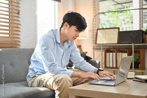 Happy young asian man browsing internet, booking hotel online on laptop while sitting on sofa at home