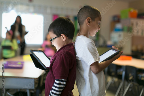 Two smart little boys practice their lessons on their respective tablets standing back-to-back at the digital interactive class.