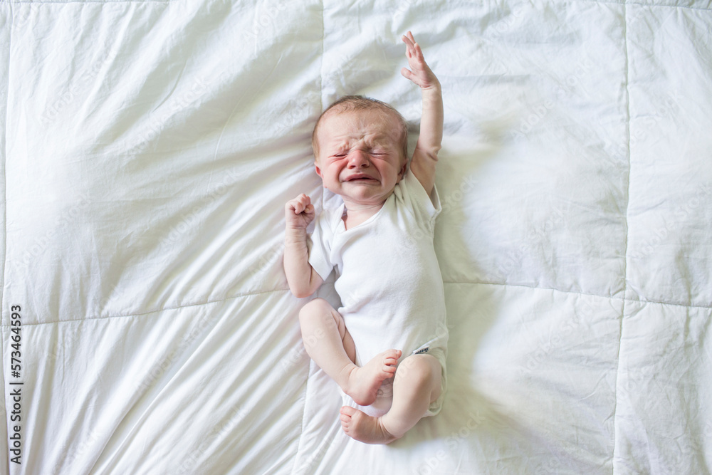 Crying Newborn Boy in White Onesie Lays on White Bed Crying Newborn Boy in White Onesie Lays on White Bed