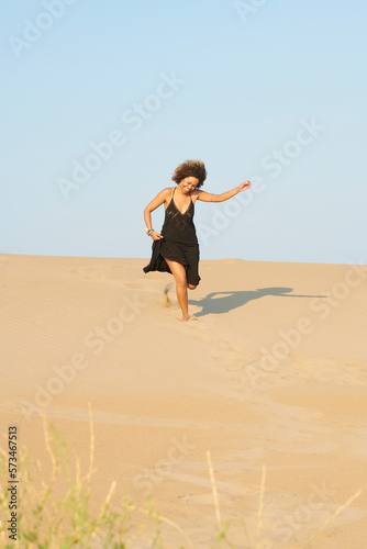 Full length portrait of cheerful woman with dark brown hair and black long summer dress running down the sandy hill happily with arm up. She is holding the skirt of dress.