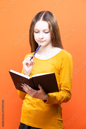 Blond Girl in Yellow Shirt Posing isolated on Orange Background as People Lifestyle Concept While Writing Notes in Notebook.