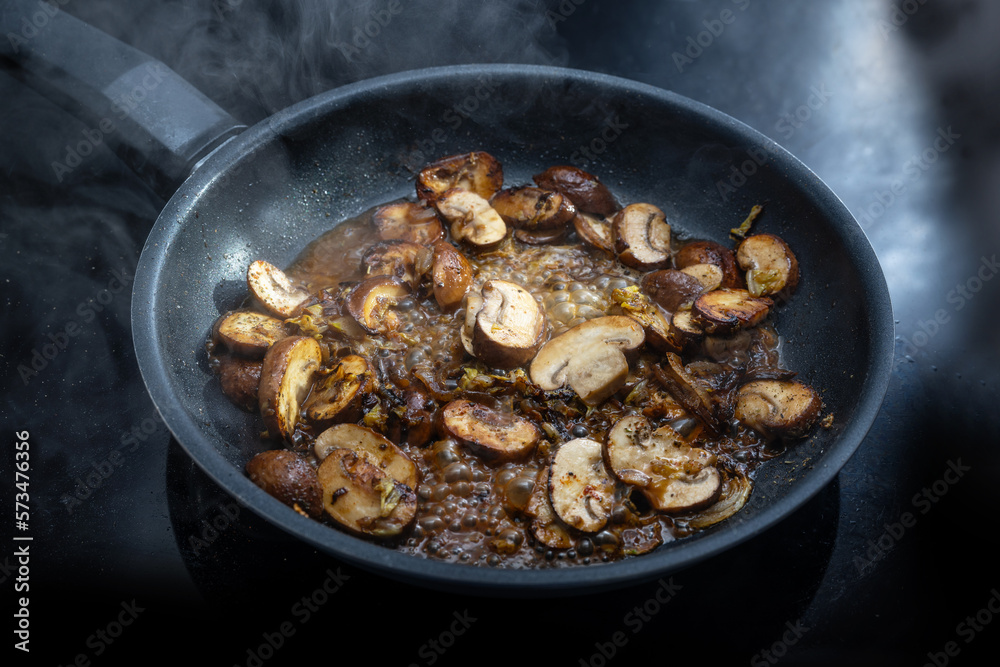Mushrooms with sauce are cooked in a steaming black frying pan on the cooktop, preparing a vegetarian meal, copy space, selected focus