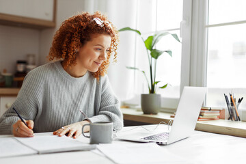 Cute university student with curly red hair doing homework sitting at kitchen table with coffee cup and wired earphones next to big window, watching online tutorial on laptop and writing in copybook