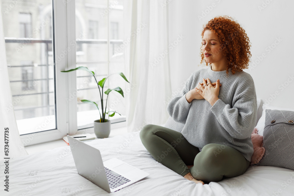 Pretty redhead plus size female sitting on bed in lotus posture with hands on chest doing pranayama techniques in front of laptop, breathing deeply, reaching inner balance after stressful day - obrazy, fototapety, plakaty 