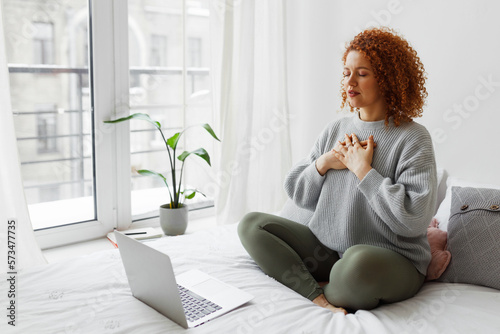 Pretty redhead plus size female sitting on bed in lotus posture with hands on chest doing pranayama techniques in front of laptop, breathing deeply, reaching inner balance after stressful day